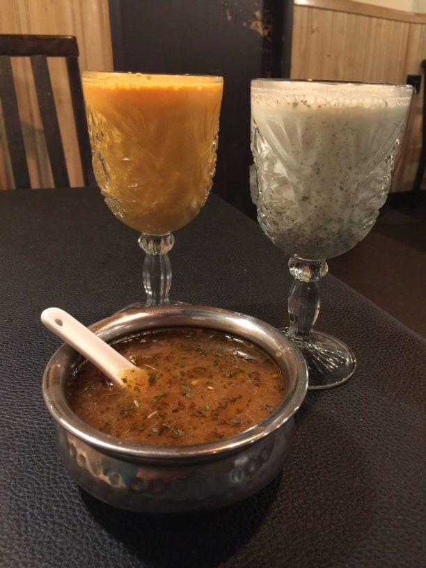This image shows doogh, a cucumber-mint yogurt drink, in a crystal goblet in the back left of the image. Pictured back right is Joose e Am, a bright orange mango yogurt drink. The two drinks are placed behind a bowl of aush, a traditional Afghan vegetable soup, in a filligreed silver dish