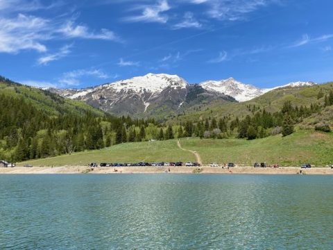 Scenic view of Tibble Fork Reservoir in American Fork Canyon, Utah, with snow-capped mountains, green hills, and a clear blue sky.