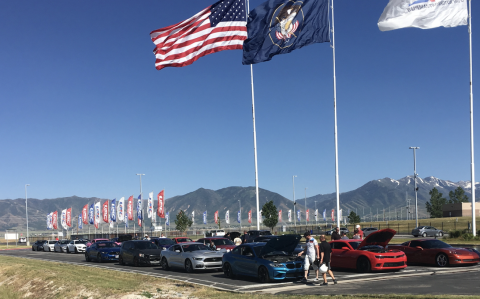 Row of performance cars parked at Utah Motorsports Campus under American and Utah state flags with mountain backdrop.