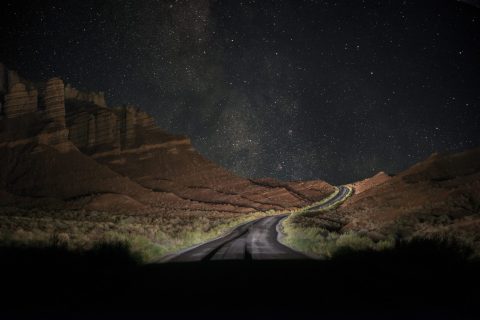 stars in the sky with a lit up road and the orange=-red rock mountains at night