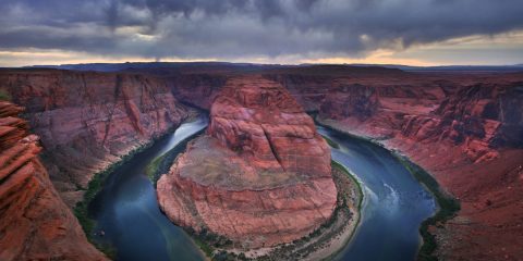 red rock horseshoe shapped canyon with a river of water and trees nerby with cloudy skies
