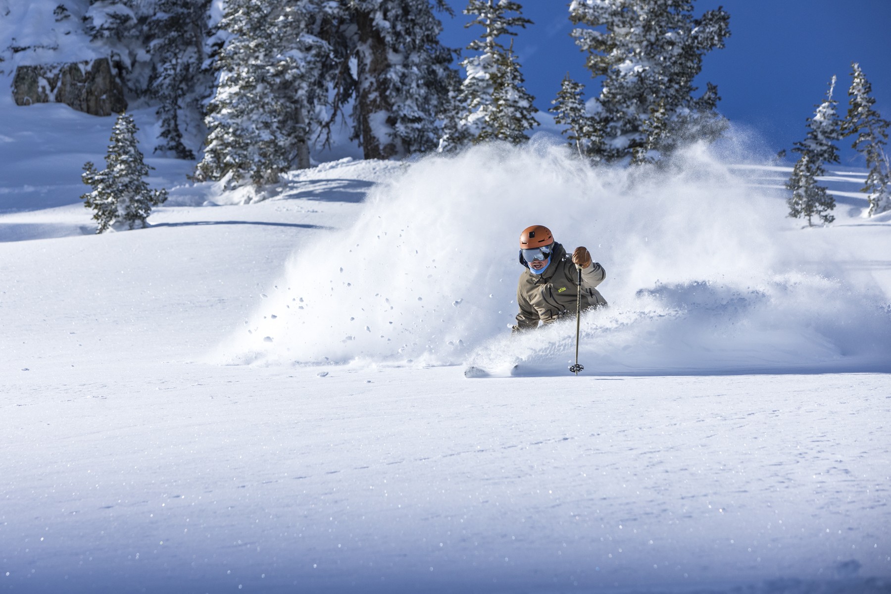 Skier carving through deep powder on a steep, snowy mountain slope with scattered trees under a clear blue sky.