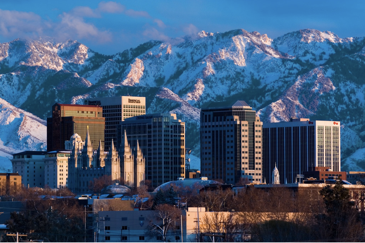 Salt Lake City skyline with downtown buildings and the Salt Lake Temple in the foreground, set against snow-covered Wasatch Mountains under a clear blue sky.