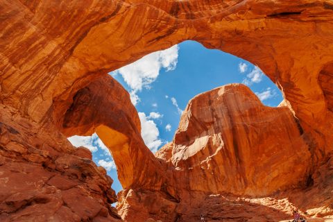 An massive arch at Arches National Park, in red sandstone with a bright blue sky and clouds above the Utah desert