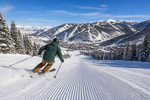 Skier carving down a freshly groomed run in Salt Lake City, overlooking a mountain village and snowy peaks.