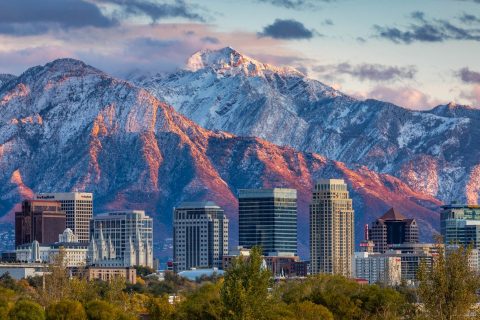 Salt Lake City skyline at sunset during the fall, showing downtown buildings positioned directly against the snow-capped Wasatch Mountains, highlighting the proximity to outdoor activities.